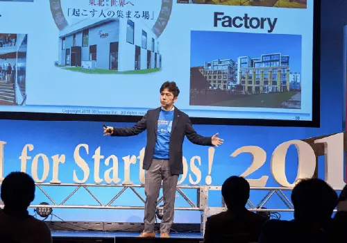 スライドを背にしてステージ上で話す男性